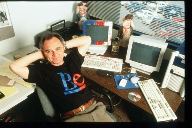 Jean-Louis Gassée seated at a desk in an office, wearing a black shirt and leaning back with hands behind his head.