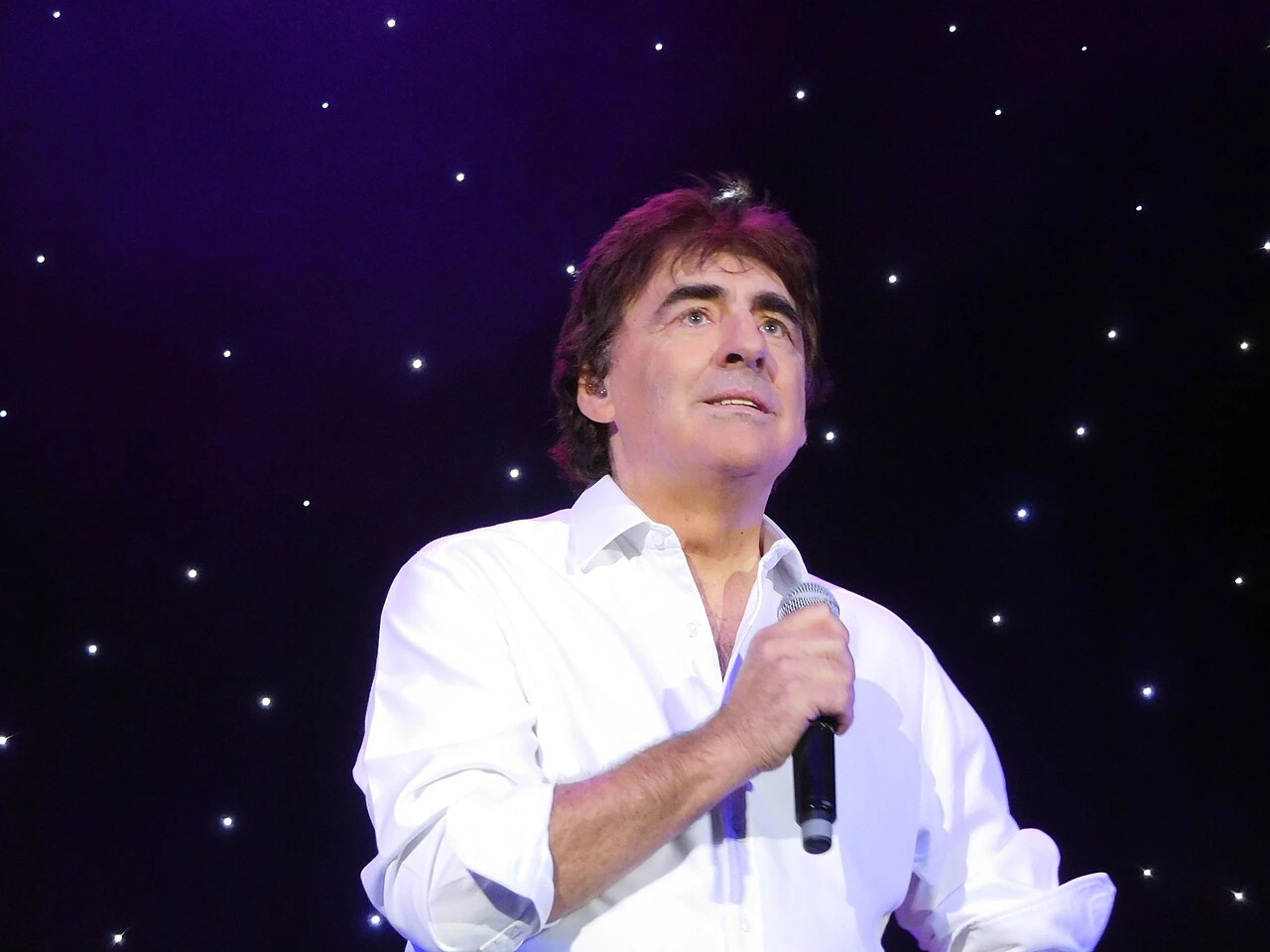 Claude Barzotti singing on stage, holding a microphone, against a starry dark background